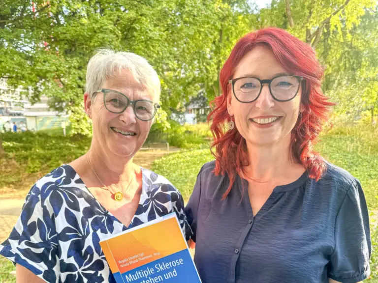 Zwei lächelnde Frauen stehen nebeneinander im Freien, eine hält ein Buch über Multiple Sklerose in den Händen.