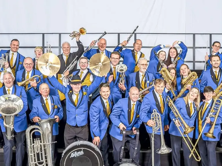 Grosse Musikkapelle in blauen Uniformjacken posiert mit Blasinstrumenten und Trommeln auf einer Bühne.