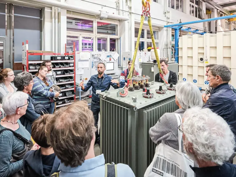 Besuchergruppe steht um einen grossen Transformator in einer Werkshalle und hört einem Mitarbeiter zu.