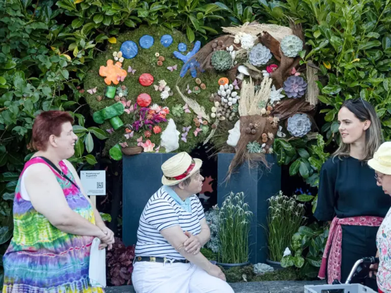 Menschen unterhalten sich vor einer mit bunten Blumen und Naturmaterialien geschmückten Hecke.