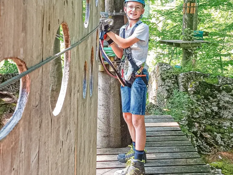 Ein Junge mit Helm und Klettergurt steht gesichert auf einer Plattform im Hochseilgarten.