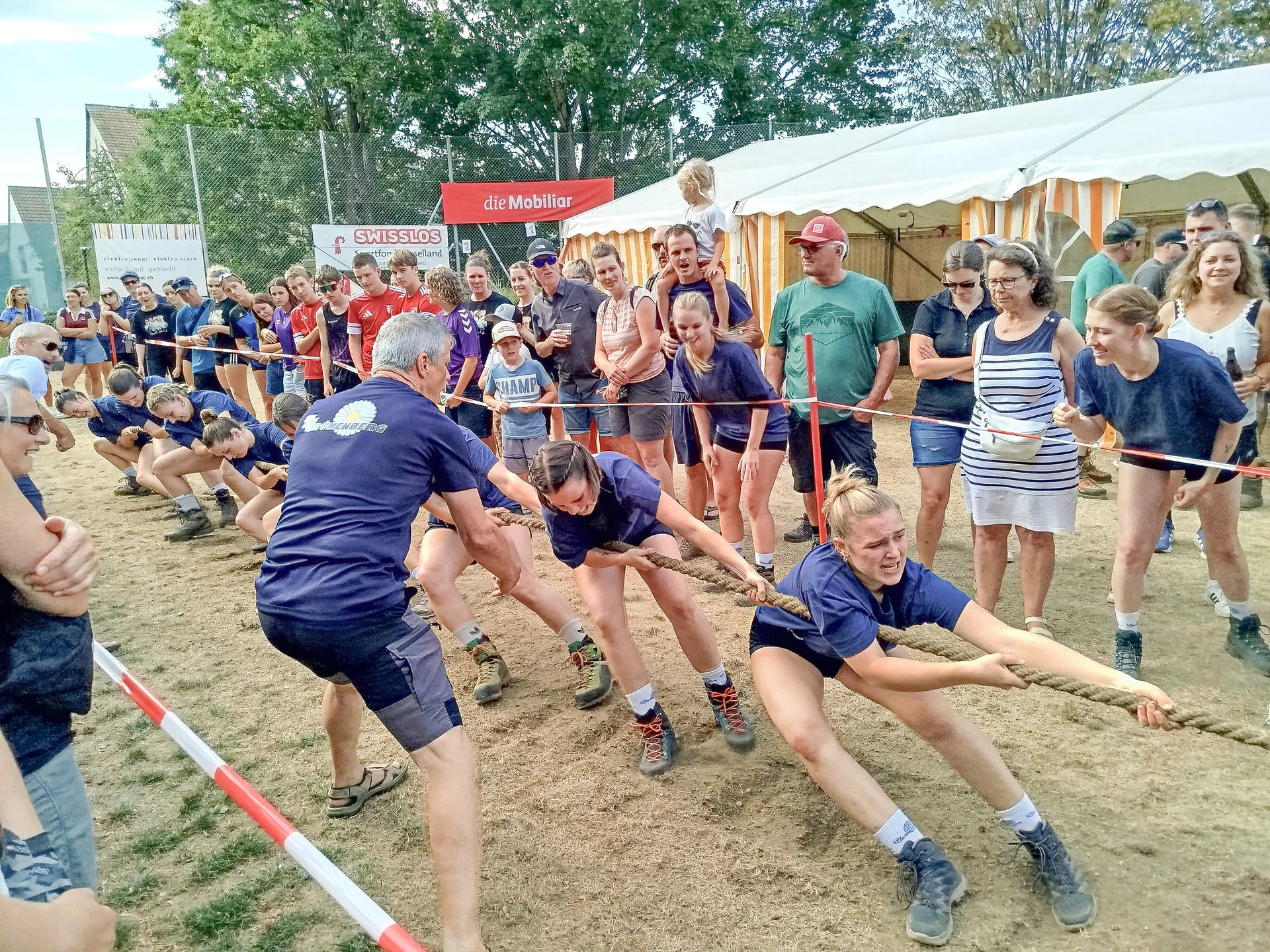 Mehrere Personen in blauen Trikots ziehen beim Tauziehen an einem Seil auf sandigem Boden.