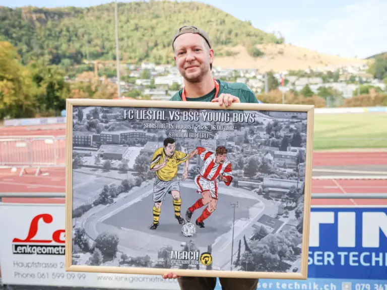 Ein Mann mit Cappy hält ein gerahmtes Bild mit zwei Fussballspielern vor einem Fussballstadion.