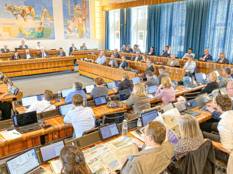 Blick in einen Ratssaal mit vielen Sitzreihen, an Pulten mit Laptops sitzen zahlreiche Menschen während einer Sitzung.