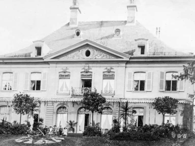 Historische Aufnahme einer herrschaftlichen Villa mit Türmchen, Fensterläden und Garten, davor stehen mehrere Personen.
