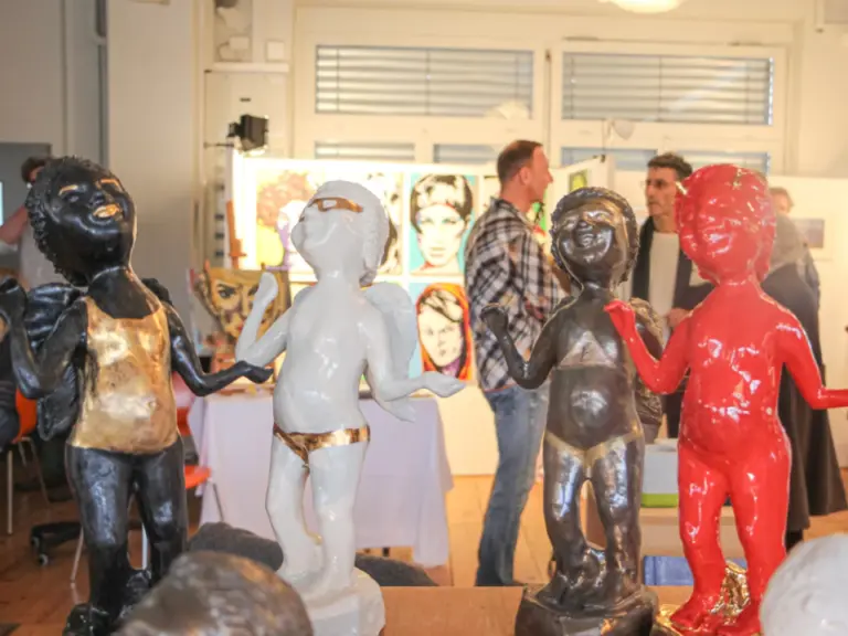 Bunte Skulpturen von Figuren im Vordergrund, im Hintergrund Besucher bei einer Kunstausstellung mit Gemälden.