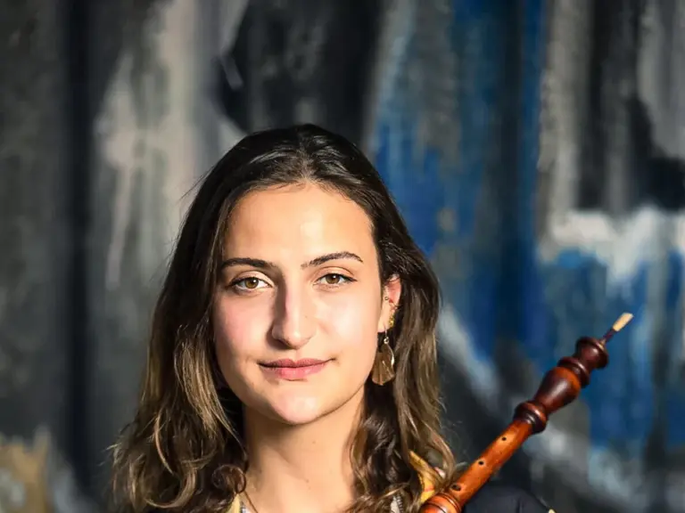 Junge Frau mit lockigem Haar hält ein Holzblasinstrument vor gemusterter Bluse, Graffiti-Wand im Hintergrund.