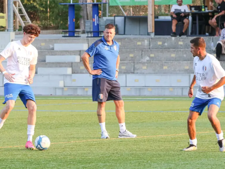 Fussballspieler und ein Trainer in Trainingskleidung auf dem Rasen, im Hintergrund Zuschauer auf einer Tribüne.