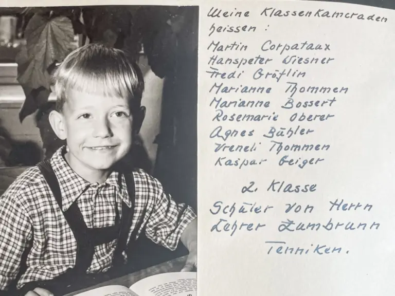 Ein lächelnder kleiner Junge im Karohemd mit Hosenträgern sitzt vor einem aufgeschlagenen Bilderbuch, daneben handgeschriebene Klassenliste.