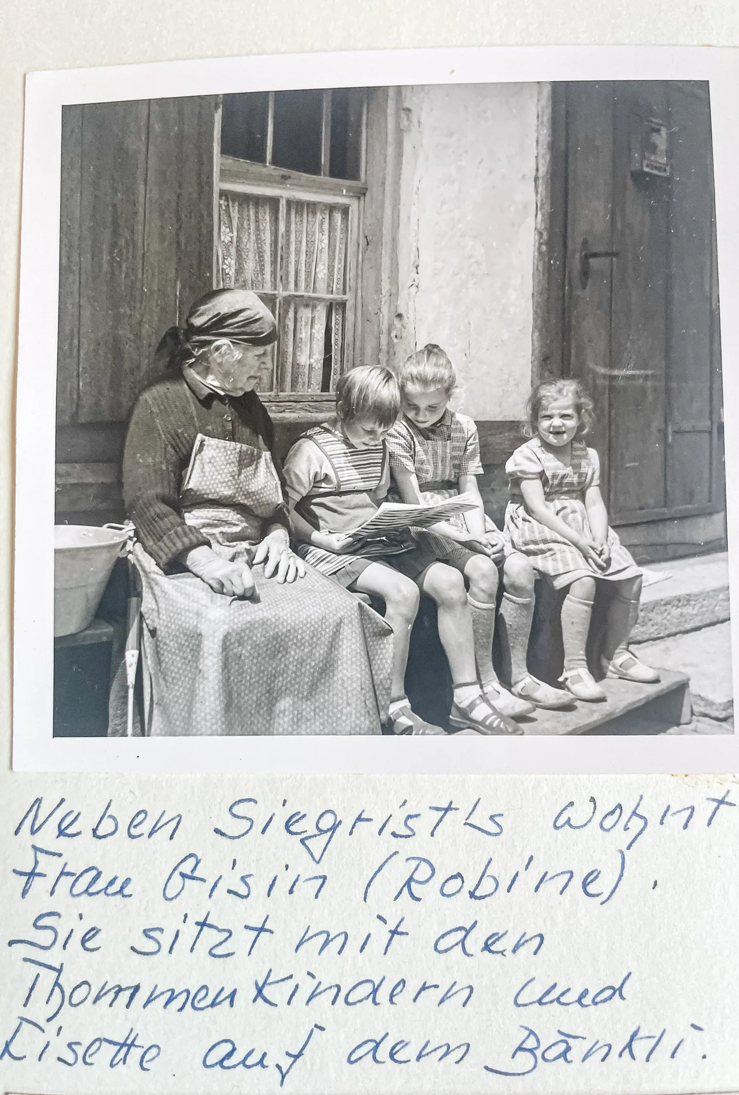 Eine ältere Frau sitzt mit drei Kindern auf einer Bank vor einem Haus und schaut in ein Buch.