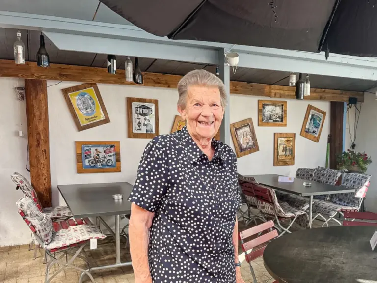 Eine ältere Frau mit gepunkteter Bluse lächelt in einer überdachten Restaurantterrasse.