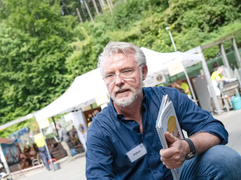 Ein Mann kniet vor einem hellen Hund mit blauem Geschirr und streichelt ihn, im Hintergrund ein Marktstand.