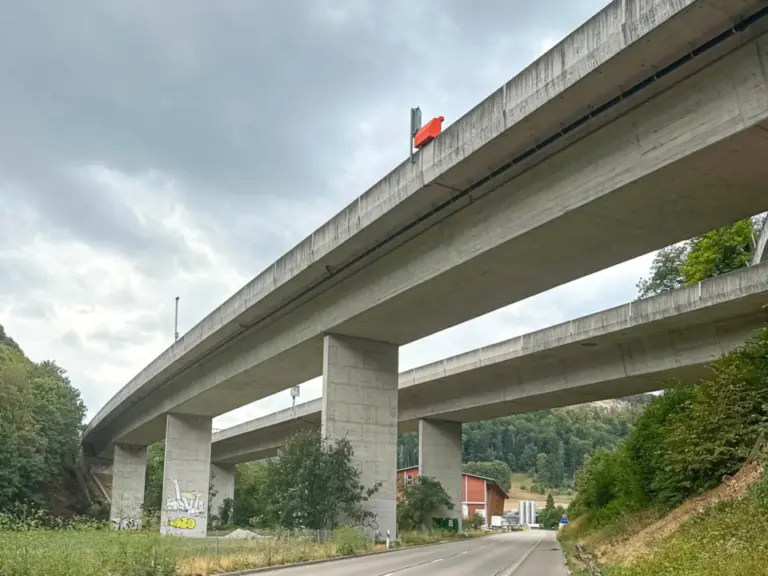 Mehrstöckige Betonautobahnbrücke überspannt eine leere Landstrasse zwischen bewaldeten Hügeln.
