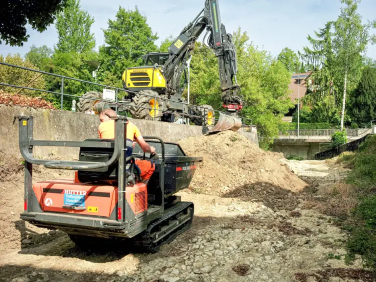 Ein Bauarbeiter sitzt auf einem Kettendumper neben einem Bagger auf einer Baustelle mit Erdaushub.