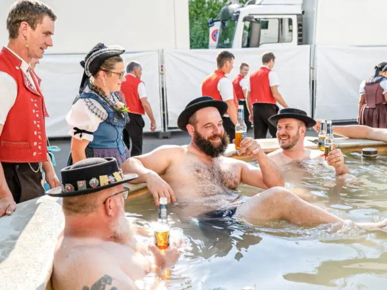 Männer mit Trachtenhüten sitzen lachend und bierselig in einem Wasserbecken, im Hintergrund Trachtenträger.