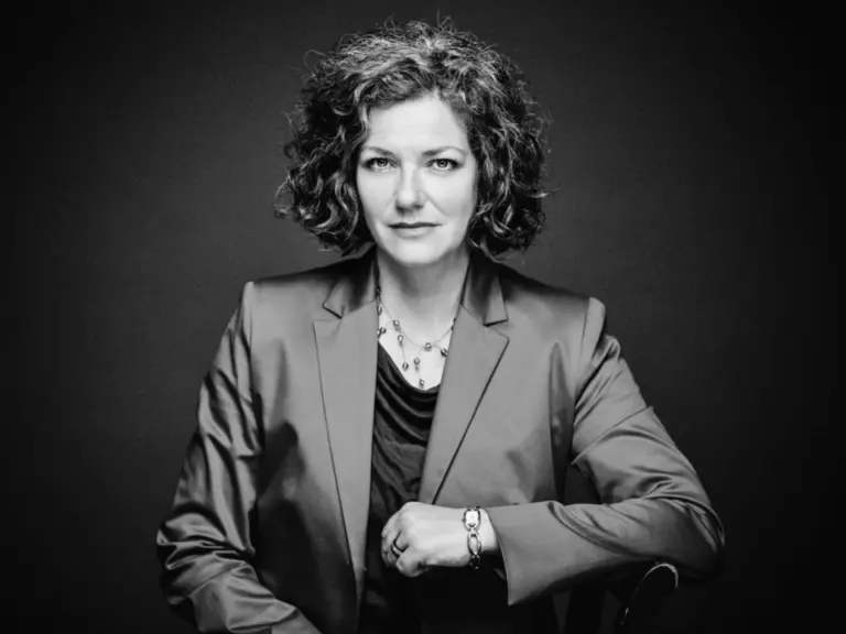 Schwarz-weiß-Porträt einer Frau mit lockigem Haar und Blazer, die in die Kamera blickt.