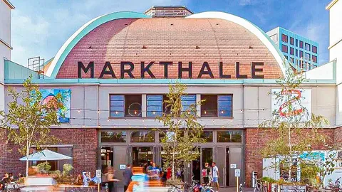 Historisches Backsteingebäude mit gewölbtem Dach und Schriftzug „Markthalle“, davor Passanten und Fahrräder.