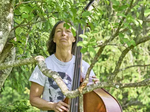 Eine Frau steht mit einem Kontrabass zwischen Bäumen auf einer grünen Wiese im Freien.