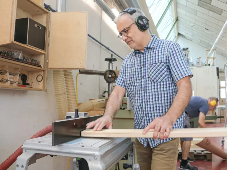 Ein Mann mit Gehörschutz und Brille führt ein Holzbrett an einer Werkstattmaschine entlang.