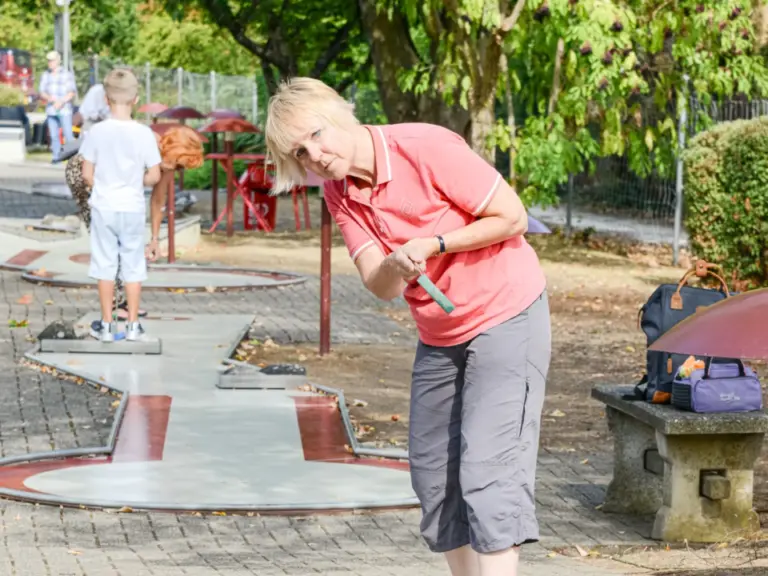 Eine Frau in rosa Poloshirt beugt sich konzentriert über einen Minigolfschläger auf einer Bahn im Freien.