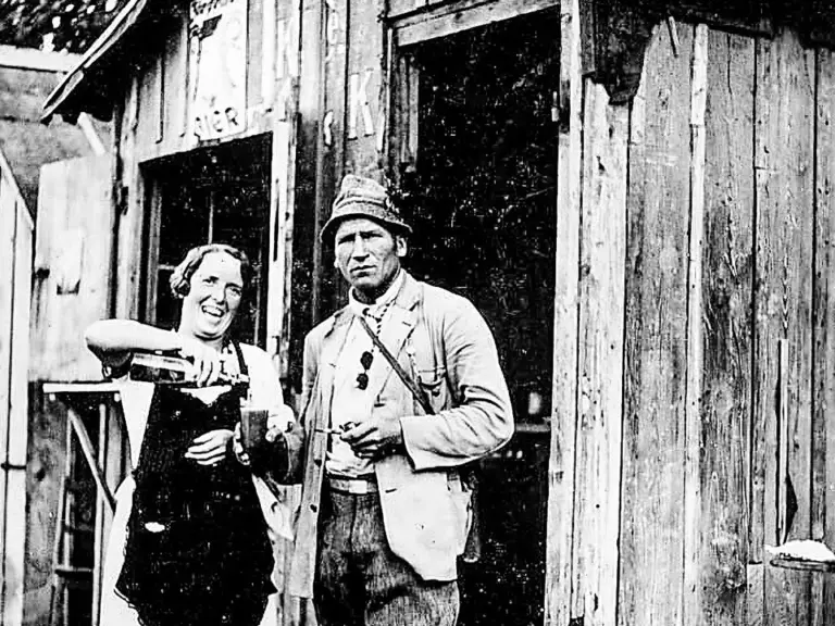 Eine Frau mit Schürze und ein Mann mit Mütze stehen vor einer Holzhütte im Wald.