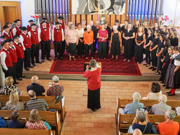 Mehrere Chöre in unterschiedlichen Trachten und Uniformen singen gemeinsam vor einer Orgel in einer Kirche, davor eine Dirigentin und Zuschauer in Bankreihen.