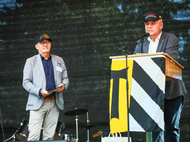 Zwei Männer mit Kappen auf einer Bühne, einer spricht am Rednerpult mit Wappen-Banner.
