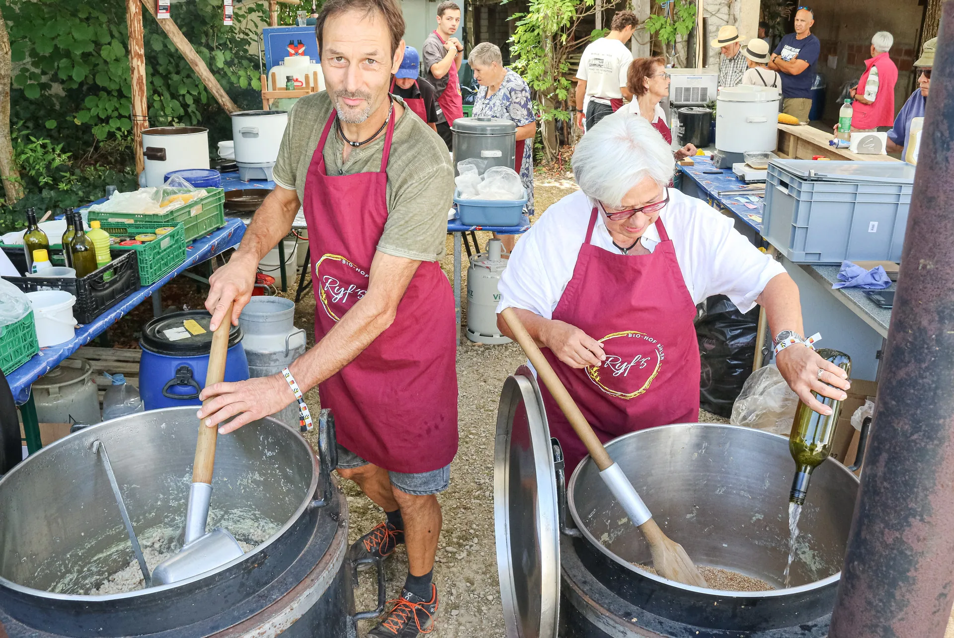 Zwei Personen in weinroten Schürzen rühren mit Holzkellen in grossen Metallbottichen im Freien.