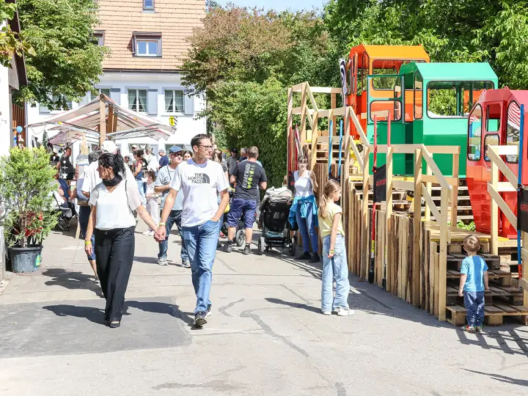 Ein Paar geht händchenhaltend über einen Platz, im Hintergrund Kinder vor bunten Holzspielgeräten.