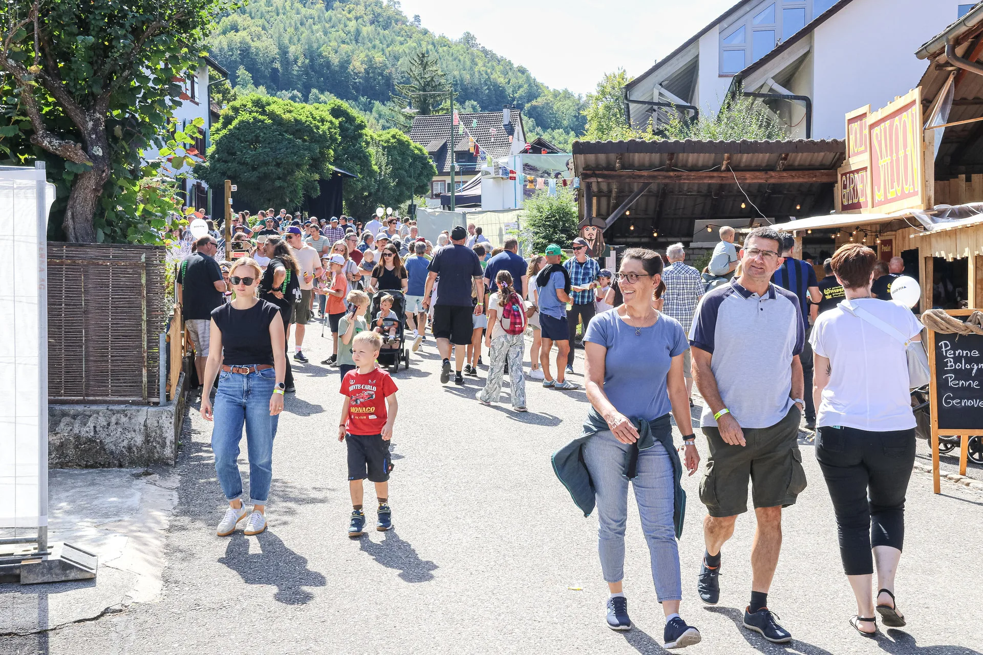 Menschenmenge schlendert an einem sonnigen Tag durch eine belebte Dorfstrasse mit Marktständen.