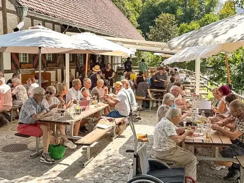 Menschen sitzen an Tischen unter Sonnenschirmen vor einem Fachwerkhaus im Freien und essen.