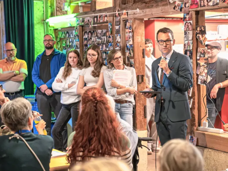 Ein Mann im Anzug spricht mit Mikrofon vor stehenden Zuhörern in einem mit Fotos dekorierten Raum.