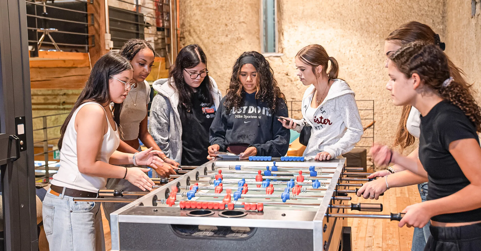 Sechs junge Frauen stehen lachend um einen Tischfussballtisch in einem rustikalen Raum.