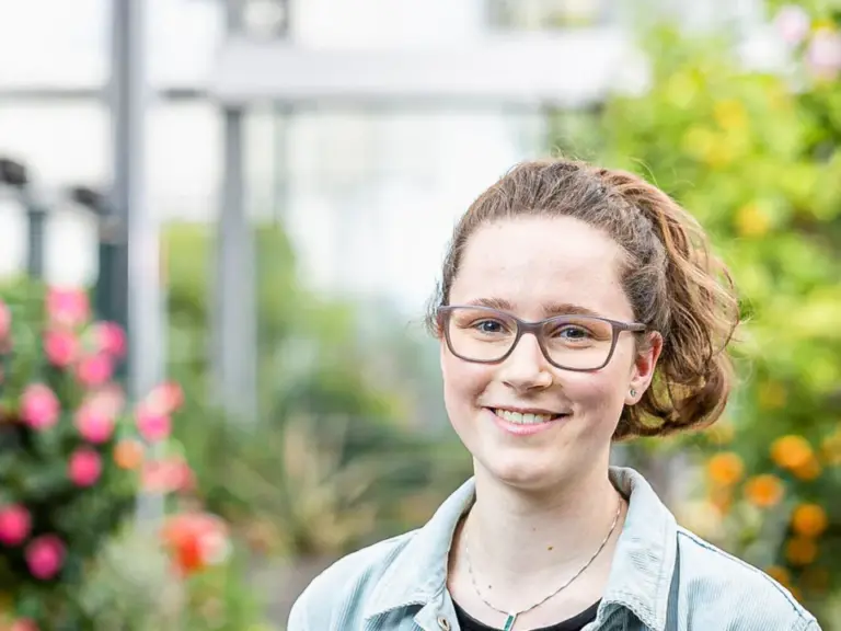 Eine junge Frau mit Brille und Jeansjacke lächelt vor blühenden Pflanzen in einem Gewächshaus.