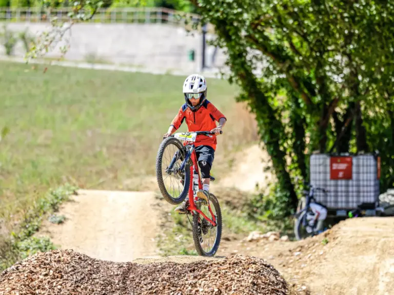 Ein Kind mit rotem Trikot und Helm springt mit dem Mountainbike über einen Erdhügel auf einer Bikepark-Strecke.