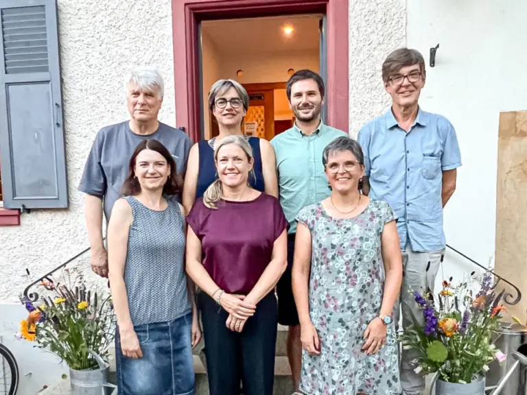 Sieben Personen stehen und knien für ein Gruppenfoto vor einem Hauseingang mit Blumensträussen.