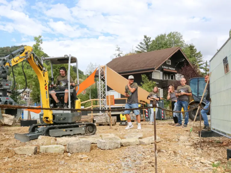Ein Mann fährt einen kleinen Bagger, mehrere Personen stehen daneben auf einer Baustelle.