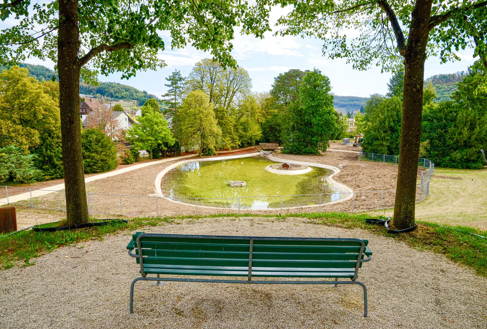 Grüne Parkbank vor einem kleinen Teich in einer Grünanlage mit Bäumen und Häusern im Hintergrund.