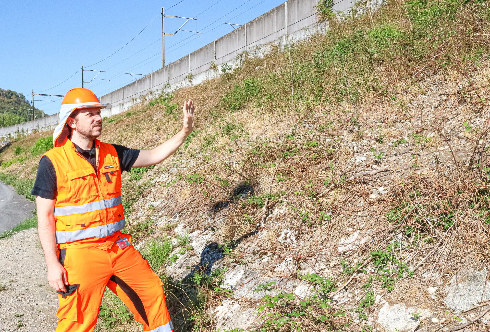 Ein Mann in oranger Arbeitskleidung und Helm steht an einer steilen, bewachsenen Böschung und deutet mit der Hand darauf.