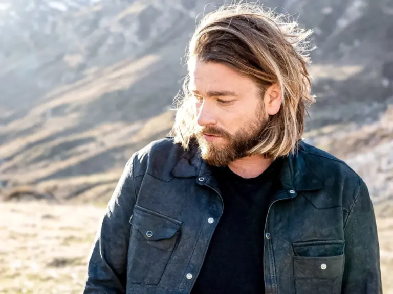 Ein bärtiger Mann mit schulterlangen Haaren blickt zur Seite, im Hintergrund eine Berglandschaft.