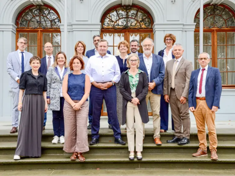 Eine Gruppe von Männern und Frauen in zwei Reihen posiert auf einer Steintreppe vor einem historischen Gebäude.
