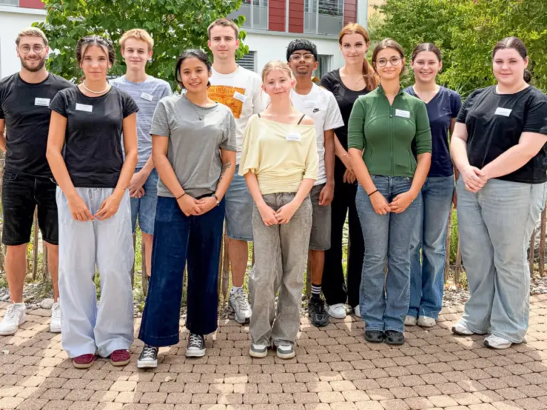 Elf junge Erwachsene stehen nebeneinander im Freien vor einem Gebäude mit Bäumen im Hintergrund.