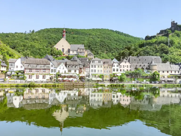 Ein Weinort mit Fachwerkhäusern und Kirche am Flussufer, im Hintergrund eine Burgruine auf bewaldetem Hügel.