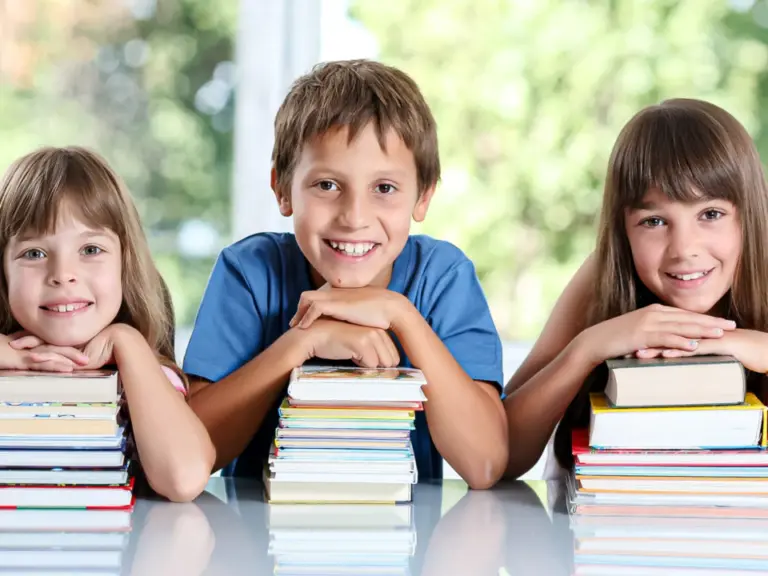 Drei lächelnde Kinder stützen sich auf Bücherstapel und blicken in die Kamera.
