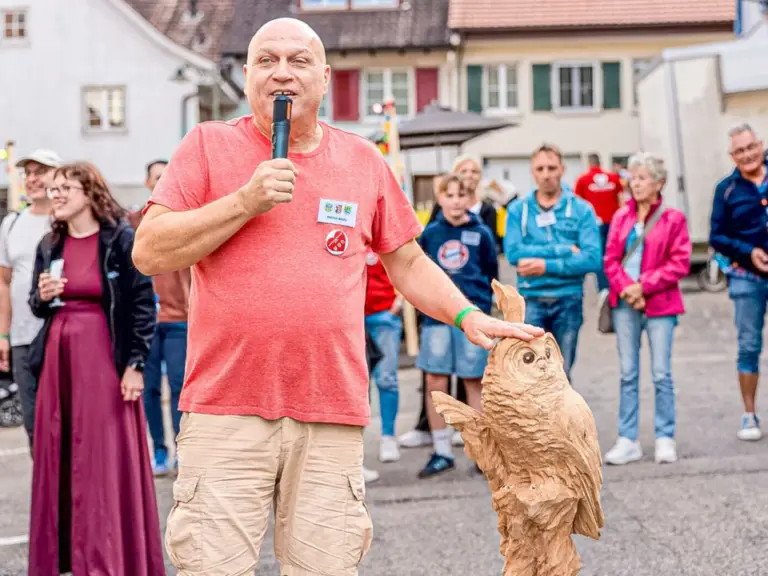 Ein Mann spricht ins Mikrofon und legt die Hand auf eine geschnitzte Holzeule vor sich.