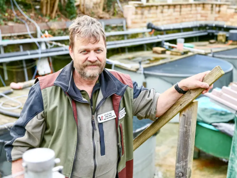 Ein Mann mittleren Alters in Arbeitsjacke lehnt an einem Holzpfosten vor grossen Wasserbecken im Freien.