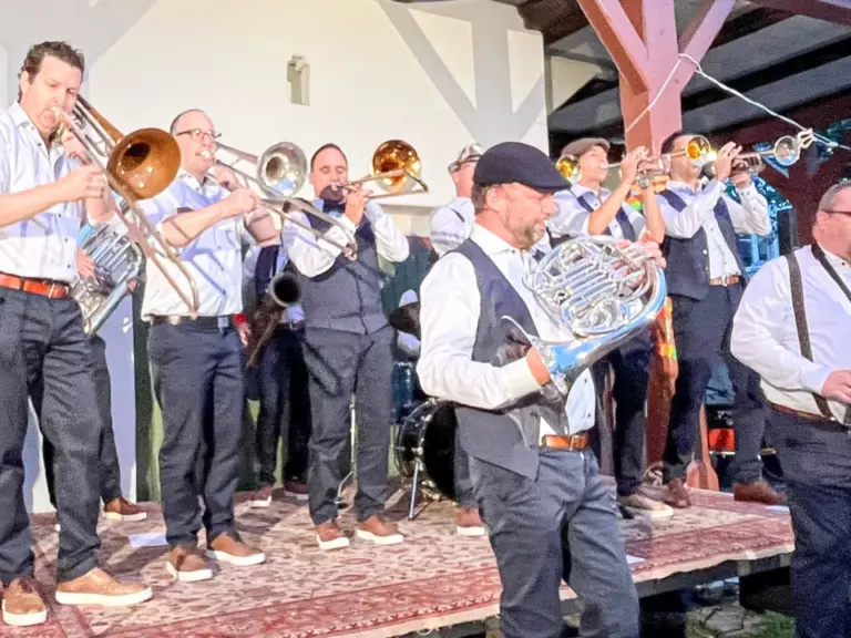 Eine Blaskapelle mit mehreren Musikern spielt auf einer erhöhten Bühne vor einem Fachwerkhaus im Freien.