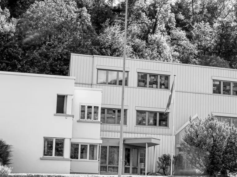 Ein modernes Firmengebäude mit Flachdach vor bewaldetem Hügel, Schwarz-Weiss-Aufnahme.