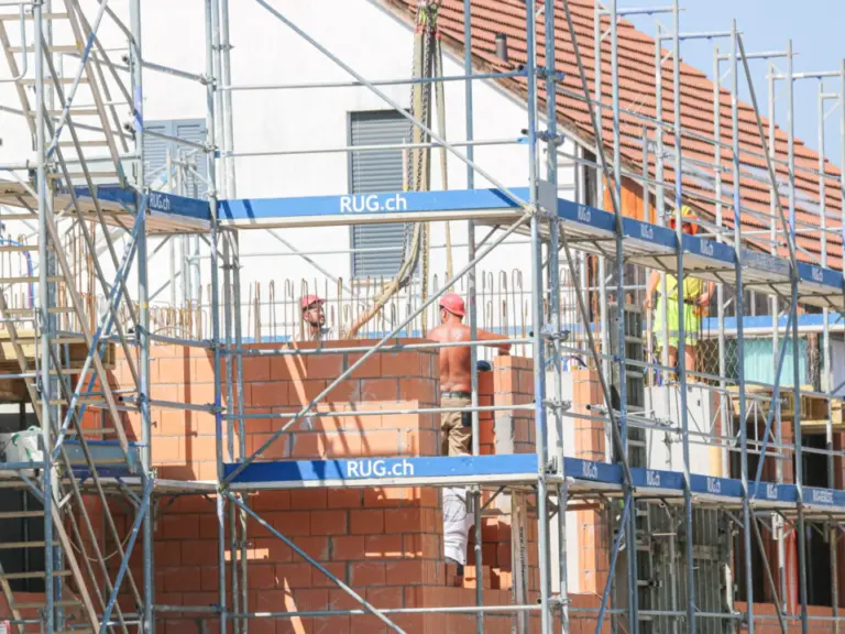 Bauarbeiter arbeiten auf Gerüsten an einem Rohbau mit Backsteinmauerwerk und Ziegeldach.