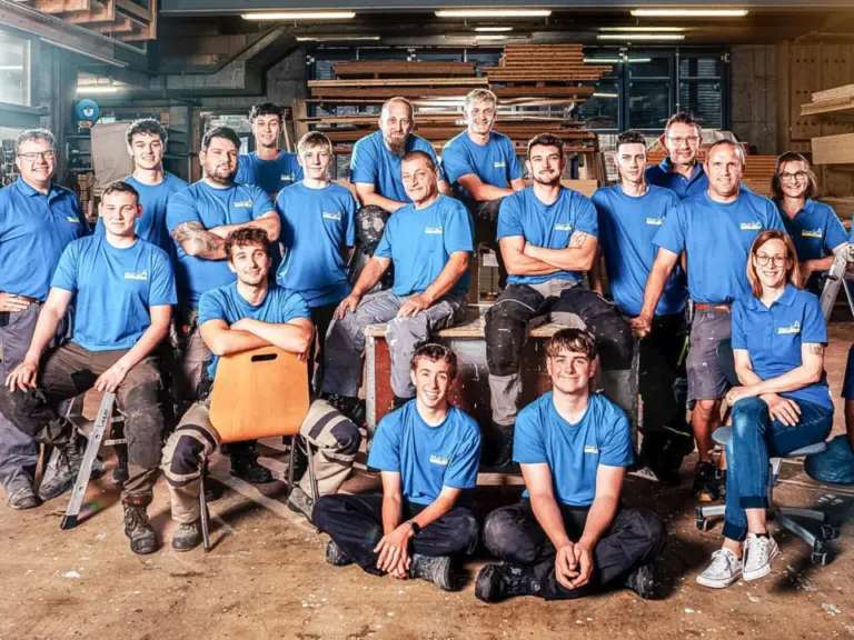 Gruppenfoto eines Teams in blauen Polo-Shirts und Arbeitshosen in einer Werkhalle einer Schreinerei.